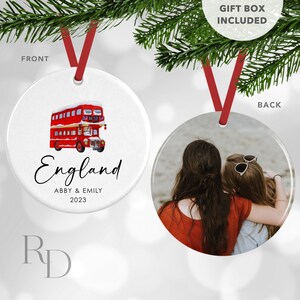 May include: A white round Christmas ornament with a red double-decker bus illustration and the text "England" with the names Abby & Emily and the year 2023. The back of the ornament features a photo of two women hugging on a beach.