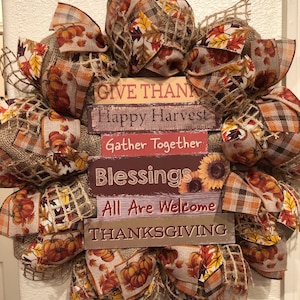 May include: A Thanksgiving wreath with burlap, plaid, and fall-themed ribbon. The wreath features a stack of wooden signs with the text "Give Thanks", "Happy Harvest", "Gather Together", "Blessings", "All Are Welcome", and "Thanksgiving".
