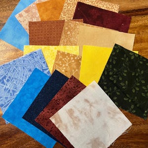 May include: A selection of fabric squares in diverse colours and patterns, including blue, yellow, brown, and green. Some squares feature floral or geometric designs, while others are solid. Suitable for quilting or craft projects.