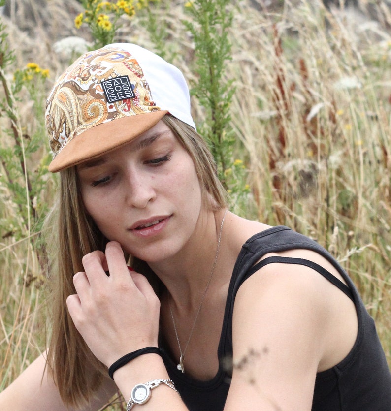 May include: A white and paisley print cap with a brown brim. The cap has a square patch with the text "SALGOSES". The cap is worn by a person in a black tank top.