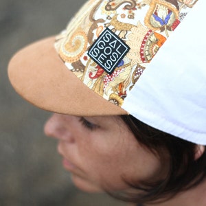May include: A baseball cap with a paisley pattern in shades of brown, gold, and red. The brim is tan, and the back is white. A black and white patch with text is on the front.