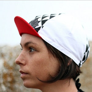 Könnte beinhalten: Eine weiße und schwarze karierte Fahrradmütze mit rotem Schirm. Die Mütze hat ein schwarz-weißes Logo mit der Aufschrift "GOAL" in einem Quadrat.