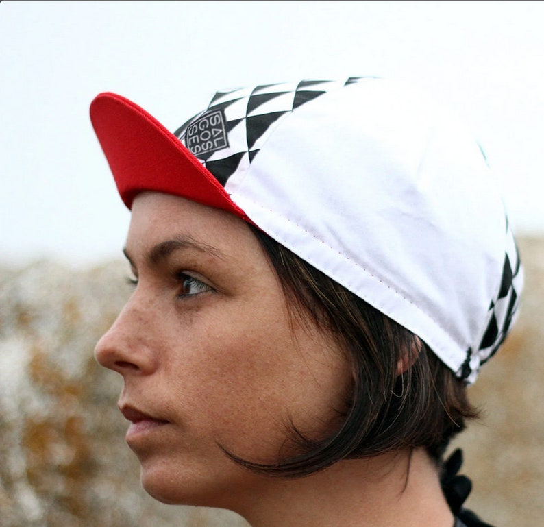Op de afbeelding: Een witte en zwarte geruite fietsmuts met een rode rand. De muts heeft een zwart-wit logo met de tekst "GOAL" in een vierkant.