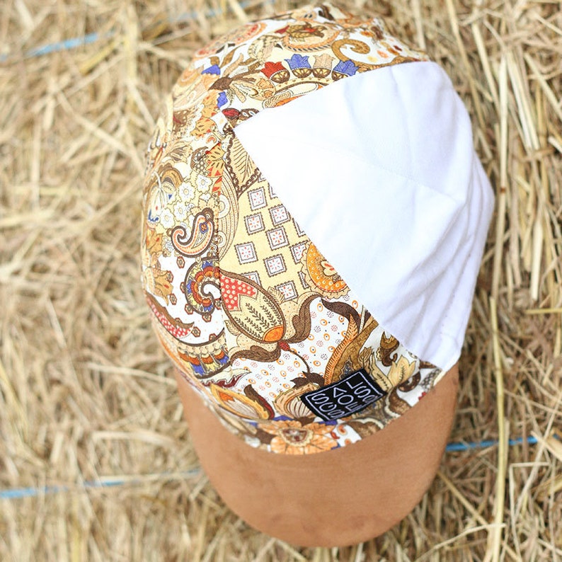 May include: A patterned cap with a brown suede brim. The cap features a mix of paisley and floral designs in shades of brown, yellow, and orange, with a white panel on the side. A small label is visible on the side.