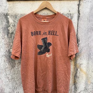 May include: A rust-colored t-shirt with the text "BORN in HELL" above a graphic of black work boots. The brand name "Danner" is printed below the boots. The shirt is hanging on a wooden hanger.