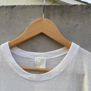 May include: A white t-shirt with a crew neck, hanging on a wooden hanger. The shirt has a tag at the back of the neck. The bottom of the shirt is not visible. The shirt appears to be made of a soft, lightweight material.