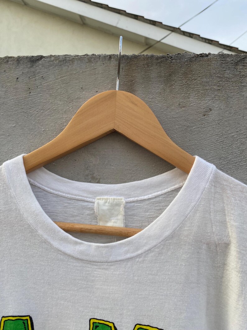 May include: A white t-shirt with a crew neck, hanging on a wooden hanger. The shirt has a tag at the back of the neck. The bottom of the shirt is not visible. The shirt appears to be made of a soft, lightweight material.