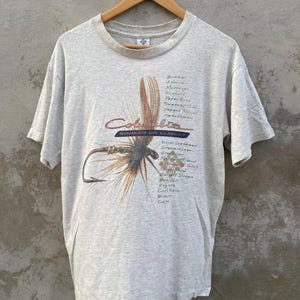 May include: A light gray Columbia t-shirt featuring a detailed graphic of a fishing fly and hook. The shirt has the text "Columbia" and "Hooked on Classics" in red and blue. Additional text is printed on the right side of the graphic.