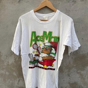 May include: White t-shirt with the text "Ace Man" in green and yellow. The graphic depicts a bulldog in a tennis outfit relaxing in a red chair with tennis balls and rackets. The shirt is hanging on a wooden hanger.