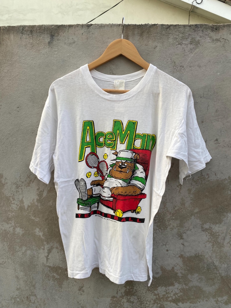 May include: White t-shirt with the text "Ace Man" in green and yellow. The graphic depicts a bulldog in a tennis outfit relaxing in a red chair with tennis balls and rackets. The shirt is hanging on a wooden hanger.