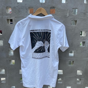 May include: White short-sleeved polo shirt with a black and white graphic on the back. The graphic includes text and an illustration of a hand. The shirt is hanging on a wooden hanger against a gray concrete wall.