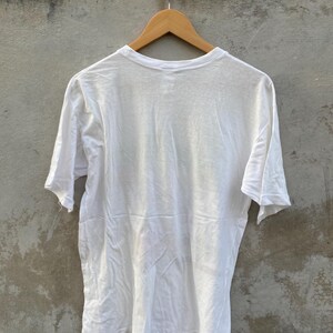 May include: A plain white t-shirt hanging on a wooden hanger. The short-sleeved shirt is made of a lightweight fabric and is slightly wrinkled. The shirt is displayed against a neutral gray background.