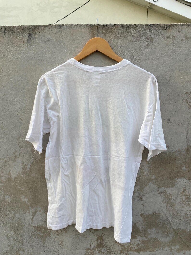 May include: A plain white t-shirt hanging on a wooden hanger. The short-sleeved shirt is made of a lightweight fabric and is slightly wrinkled. The shirt is displayed against a neutral gray background.