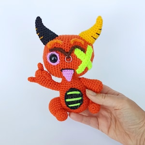 May include: Crocheted orange devil doll with black and yellow horns, a neon green cross-shaped eye patch, and a pink tongue sticking out. The doll has a black and green striped belly and is giving a hand gesture.