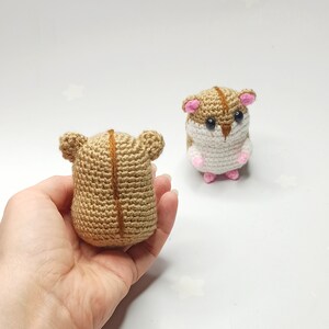 以下が含まれることがあります： 2つの編みぐるみハムスターの玩具。1つは茶色と白で、もう1つは茶色。茶色と白のハムスターは白い表面に立っています。茶色のハムスターは手で持たれています。