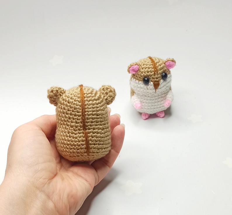 以下が含まれることがあります： 2つの編みぐるみハムスターの玩具。1つは茶色と白で、もう1つは茶色。茶色と白のハムスターは白い表面に立っています。茶色のハムスターは手で持たれています。