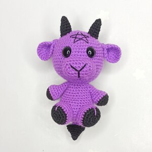 Peut inclure: Peluche de ch&egrave;vre en crochet violet avec des cornes noires, un pentagramme sur le front et des yeux noirs. La ch&egrave;vre a une petite queue noire et des sabots noirs.