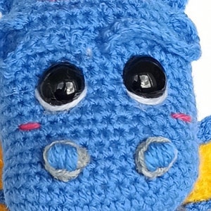 May include: A blue crocheted stuffed toy dragon with large black eyes and pink cheeks. The dragon has yellow wings and a light blue crocheted nose.