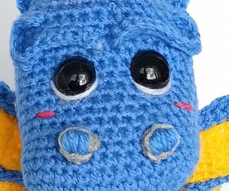 May include: A blue crocheted stuffed toy dragon with large black eyes and pink cheeks. The dragon has yellow wings and a light blue crocheted nose.