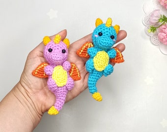 May include: Two handmade crochet dragons in a hand. One is purple with yellow and orange accents, the other is blue with yellow and orange accents. Both have small wings, horns, and a yellow belly. The dragons are small, cute, and whimsical.