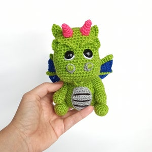 May include: A green crocheted dragon toy with pink horns, blue wings, and black eyes. The dragon is sitting on a hand.