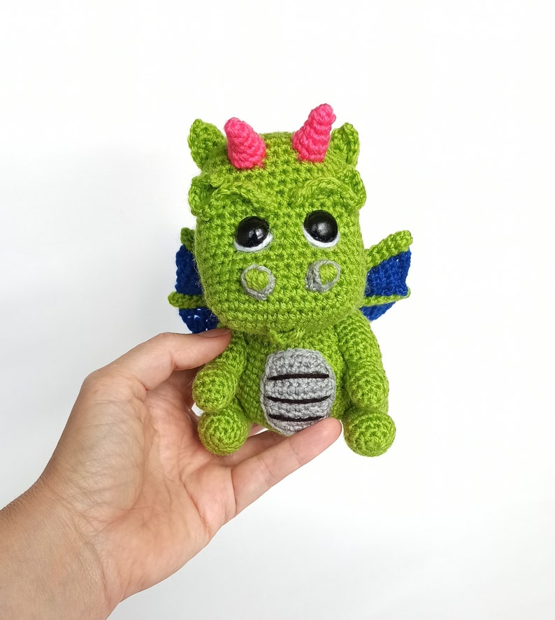 May include: A green crocheted dragon toy with pink horns, blue wings, and black eyes. The dragon is sitting on a hand.