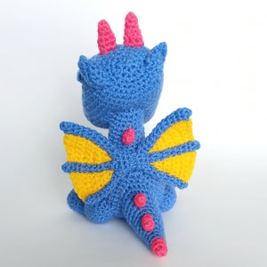 May include: A blue crocheted dragon with yellow wings and pink accents. The dragon has a long tail and is sitting on a white background.