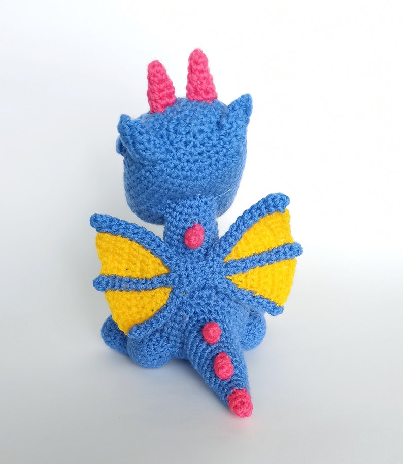 May include: A blue crocheted dragon with yellow wings and pink accents. The dragon has a long tail and is sitting on a white background.