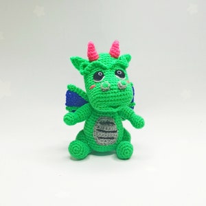May include: A green crocheted dragon toy with pink horns, blue wings, and black eyes. The dragon has a gray striped belly and is sitting on a white background.