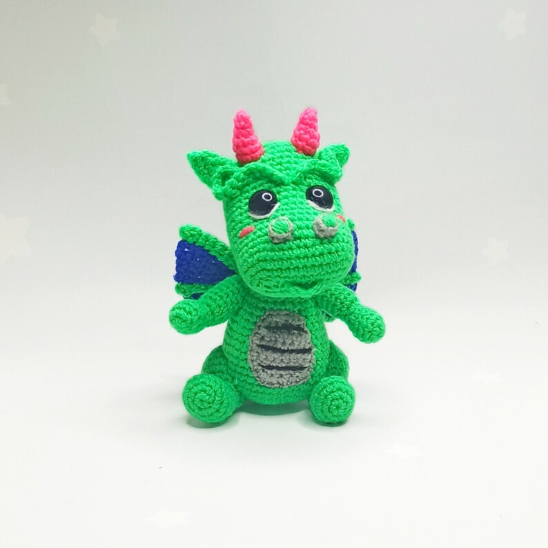 May include: A green crocheted dragon toy with pink horns, blue wings, and black eyes. The dragon has a gray striped belly and is sitting on a white background.