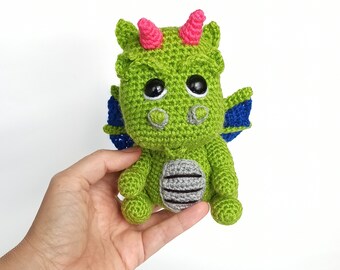 Puede incluir: Un juguete de dragón de crochet verde con cuernos rosas, alas azules y ojos negros. El dragón está sentado en una mano.