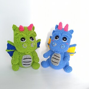 May include: Two crocheted dragon toys, one green and one blue, with pink and yellow accents. The dragons have big eyes and are sitting on a white background.