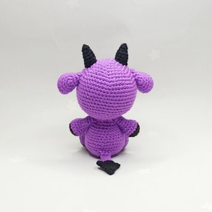 Peut inclure: Jouet en peluche crochet&eacute; violet avec des cornes et une queue noires. Le jouet est assis sur une surface blanche.