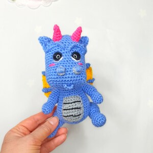 May include: A blue crocheted dragon with yellow wings, pink horns, and black eyes. The dragon has a gray crocheted belly.