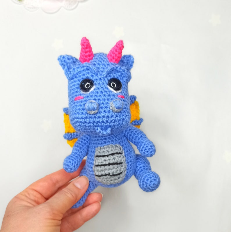 May include: A blue crocheted dragon with yellow wings, pink horns, and black eyes. The dragon has a gray crocheted belly.