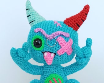 Puede incluir: Un juguete monstruo de crochet azul con detalles en rosa, verde y morado. El monstruo tiene un ojo, una lengua rosa que sobresale y un parche rosa sobre su otro ojo. El monstruo tiene dos cuernos, uno rojo y uno verde.