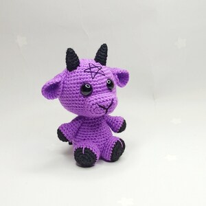 Peut inclure: Peluche de ch&egrave;vre en crochet violet avec des cornes et des yeux noirs. La ch&egrave;vre a un pentagramme cousu sur son front.