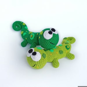 Può includere: Due giocattoli di camaleonte di uncinetto verdi con grandi occhi bianchi e un'espressione sorridente. Il camaleonte in basso è sdraiato sulla pancia con le gambe distese. Il camaleonte in alto è rannicchiato con la coda avvolta attorno al suo corpo.