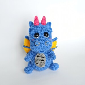 May include: A blue crocheted dragon with yellow wings and pink horns. The dragon has black eyes and a gray belly.