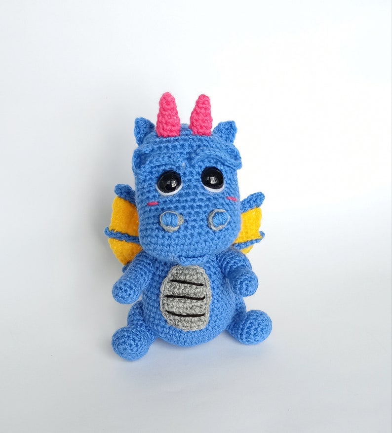 May include: A blue crocheted dragon with yellow wings and pink horns. The dragon has black eyes and a gray belly.