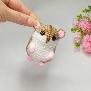 以下が含まれることがあります： 茶色と白の毛並みの小さな編みぐるみハムスターの玩具。ピンクの耳と足、黒い目です。