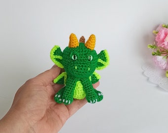 Puede incluir: Un dragón de crochet verde con cuernos amarillos y ojos verdes. El dragón está sentado en una mano.