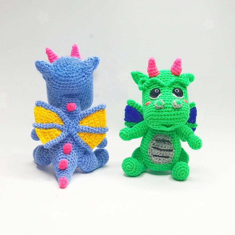 May include: Two crocheted dragon toys. One is blue with yellow wings and pink accents. The other is green with blue wings and pink accents.