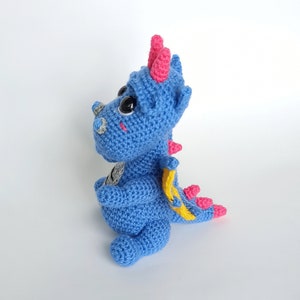 May include: A blue crocheted dragon toy with pink horns and a yellow tail. The dragon has large black eyes and a friendly expression.