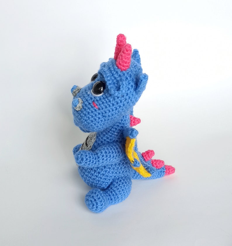 May include: A blue crocheted dragon toy with pink horns and a yellow tail. The dragon has large black eyes and a friendly expression.