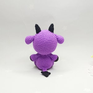 Peut inclure: Peluche en crochet violet avec des cornes et une queue noires. Le jouet est assis sur une surface blanche.