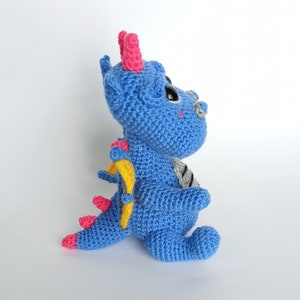 May include: A blue crocheted dragon with pink horns, yellow wings, and black eyes. The dragon is sitting on a white surface.