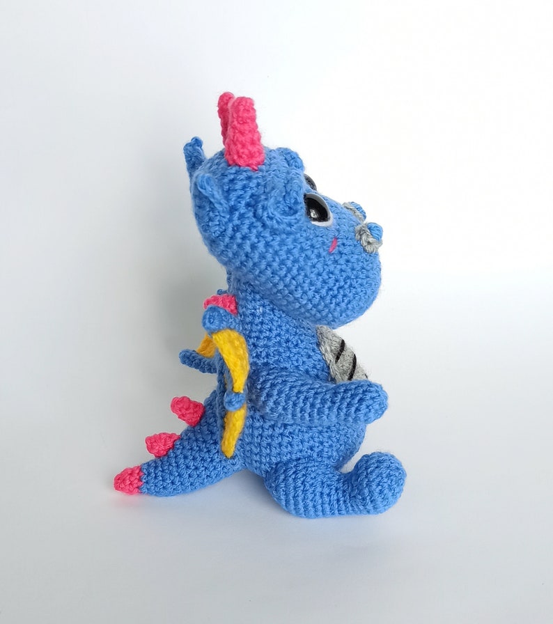 May include: A blue crocheted dragon with pink horns, yellow wings, and black eyes. The dragon is sitting on a white surface.