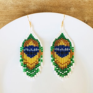 Könnte beinhalten: Ein Paar Perlenohrringe mit Federdesign. Die Ohrringe haben einen weißen Rand, grüne, goldene und blaue Perlen. Die Ohrringe hängen an goldfarbenen Haken. Die Ohrringe werden auf einem weißen Teller präsentiert.