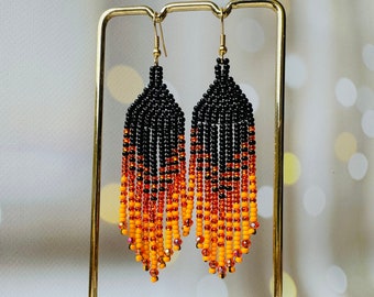 May include: A pair of long, beaded earrings with a gradient of black, orange, and yellow beads. The earrings are hanging from a gold stand.